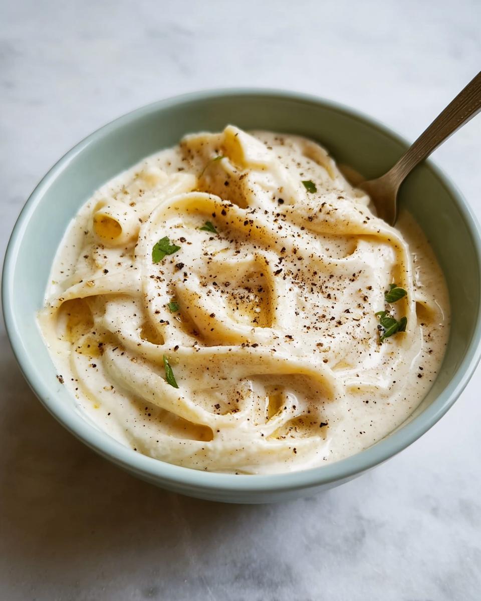 A close-up of thick, creamy Best Alfredo Sauce Recipe Ever mixed with pasta, topped with cracked black pepper and parsley.