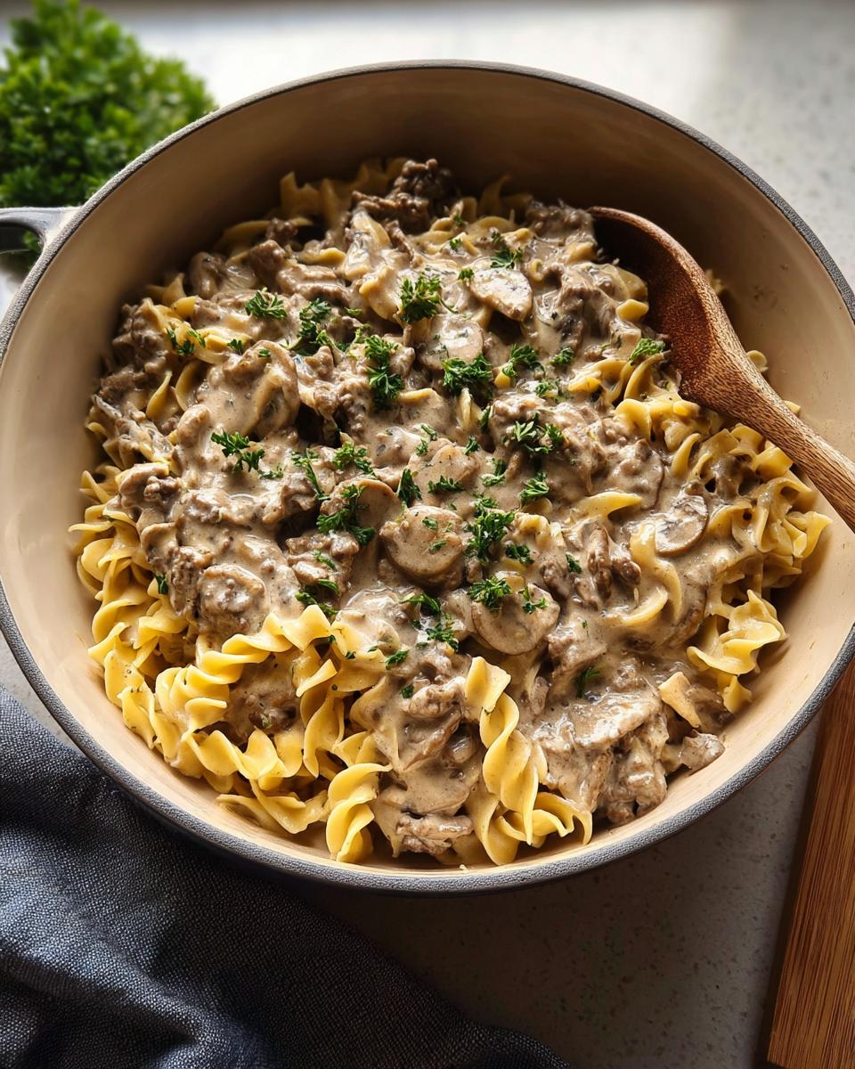 A close-up of creamy Ground Beef Stroganoff served over egg noodles in a beige bowl, garnished with parsley.