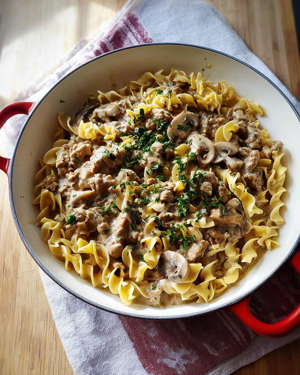 A close-up overhead view of Creamy Turkey Stroganoff served over wide egg noodles in a red-handled skillet, garnished with parsley.
