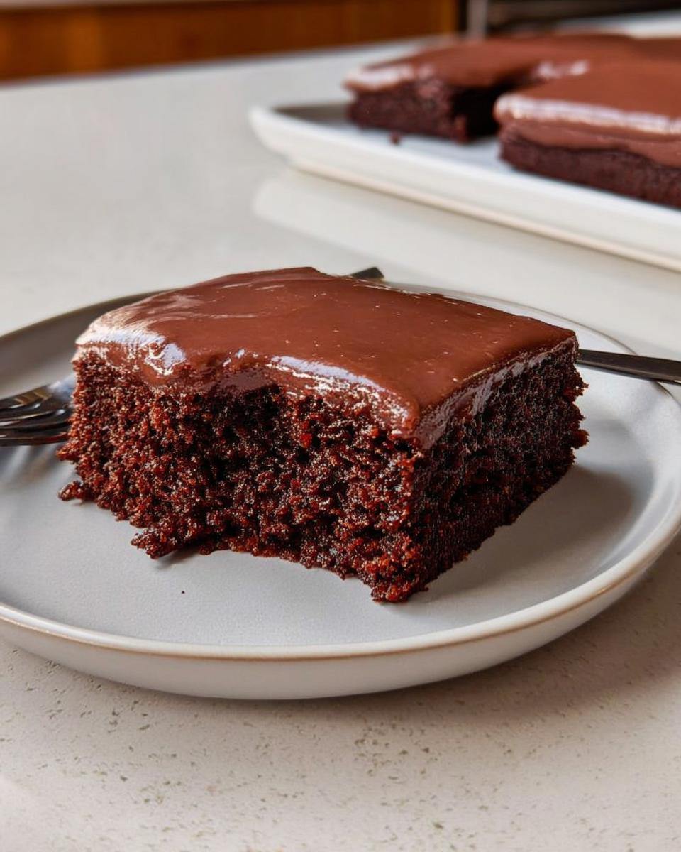 A square slice of moist, dark chocolate Easy Snacking Sheet Cake topped with glossy chocolate frosting on a light gray plate.