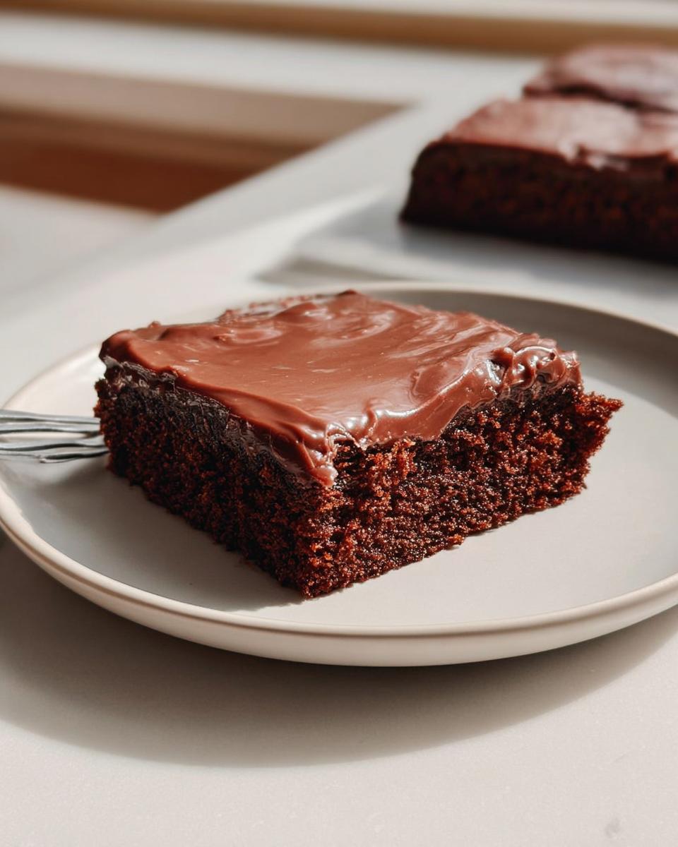 A close-up of a rich, dark slice of Easy Snacking Sheet Cake topped with glossy chocolate frosting on a light plate.