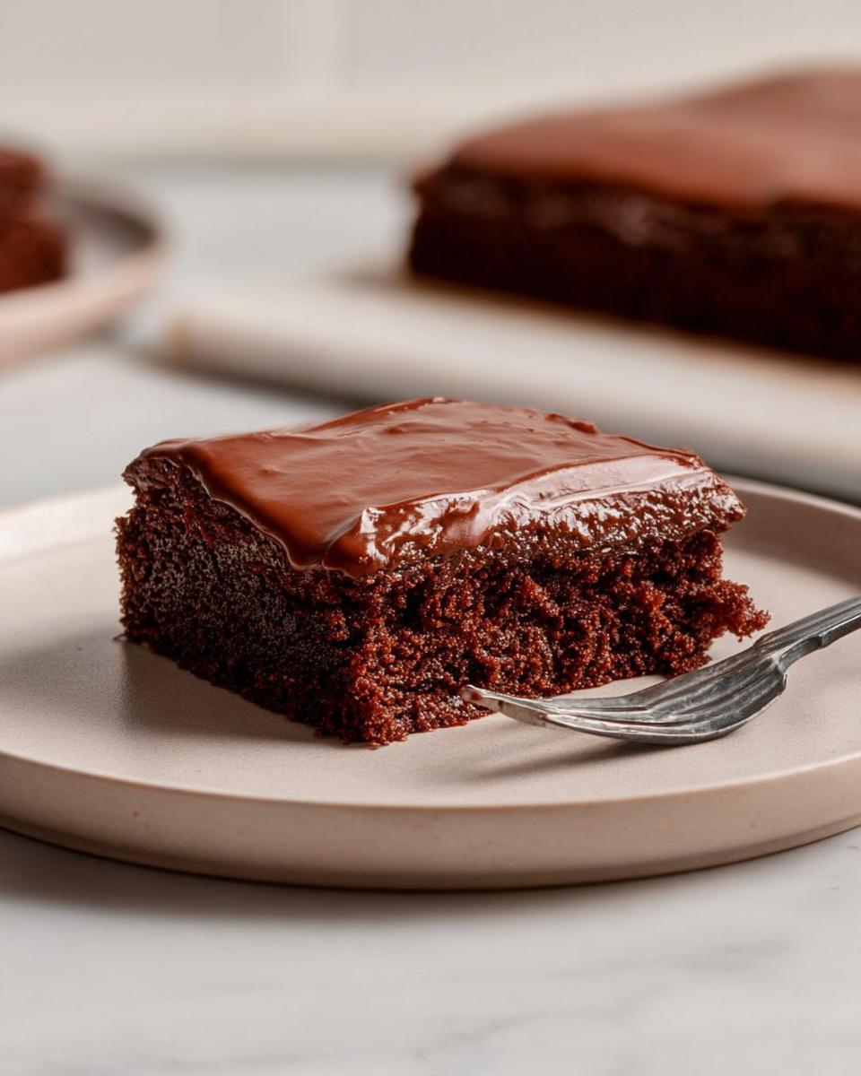 A close-up of a square slice of moist chocolate Easy Snacking Sheet Cake topped with glossy chocolate frosting, resting on a plate with a fork.