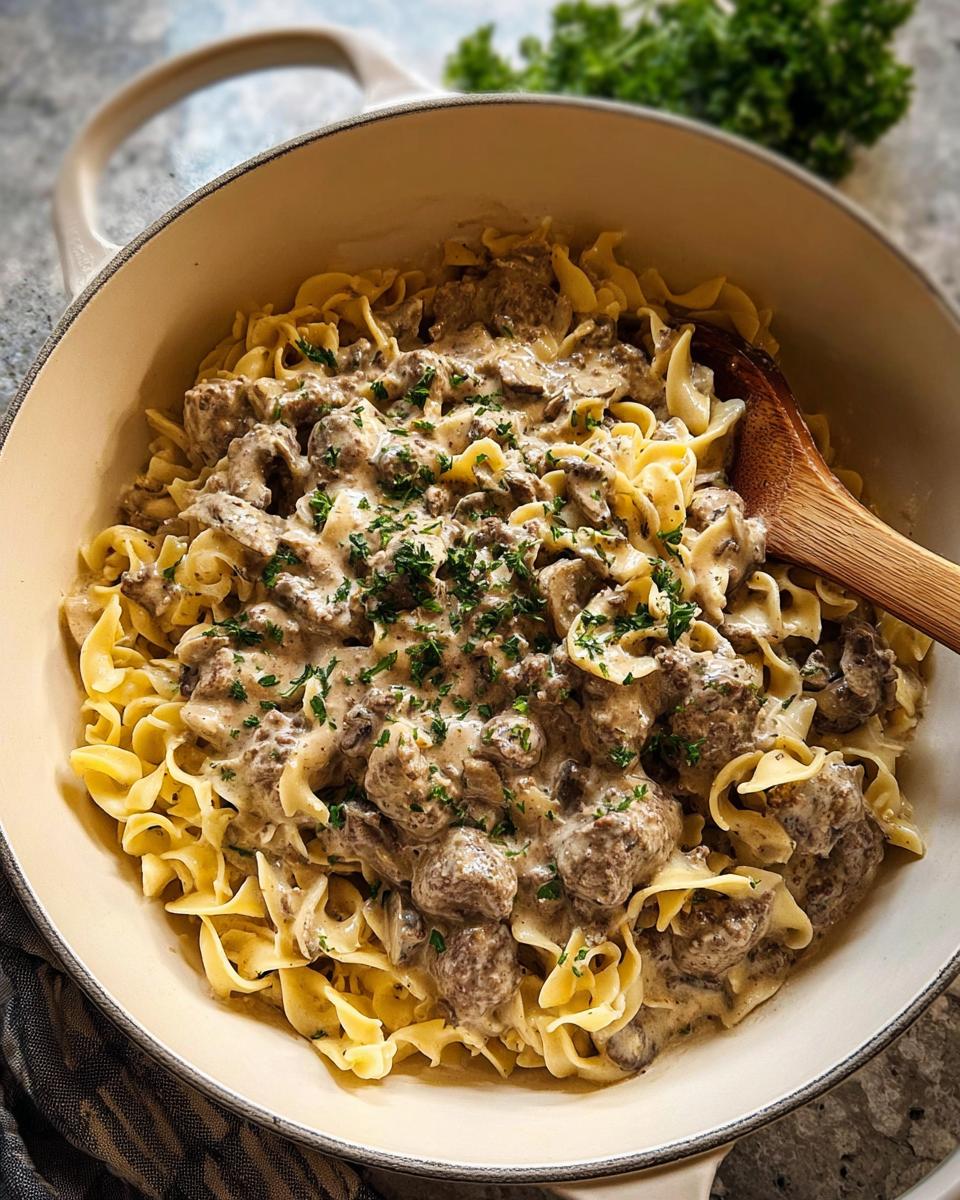 A creamy Ground Beef Stroganoff mixed with wide egg noodles, topped with fresh parsley, served in a light-colored Dutch oven.
