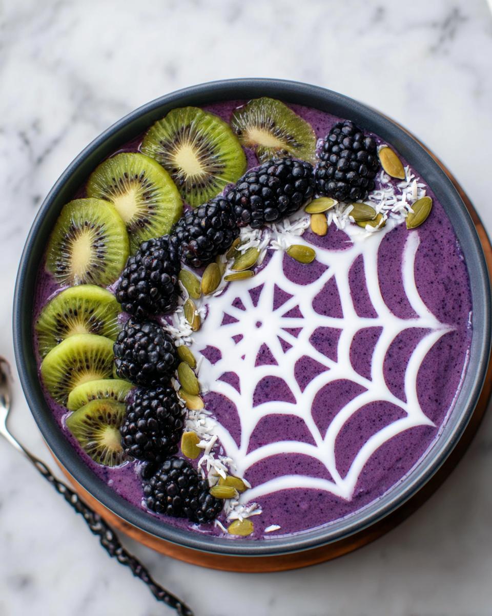 A vibrant purple Halloween smoothie bowl topped with kiwi slices, blackberries, pumpkin seeds, coconut, and a white spiderweb design.