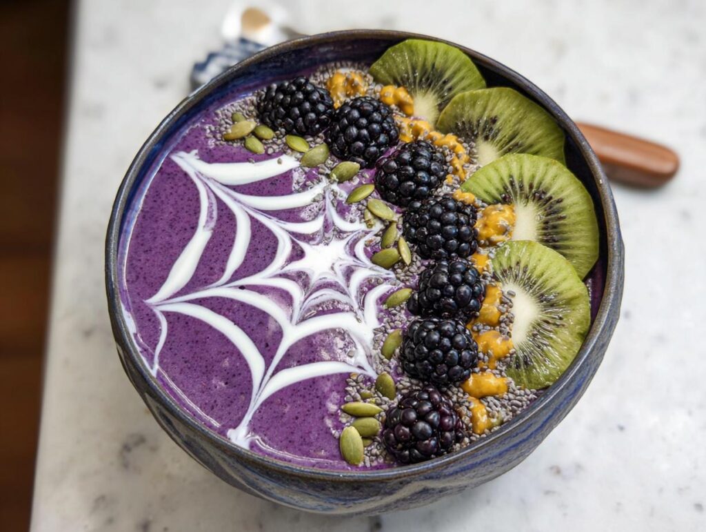 A vibrant purple Halloween smoothie bowl topped with blackberries, kiwi slices, chia seeds, pumpkin seeds, and a white yogurt spiderweb.