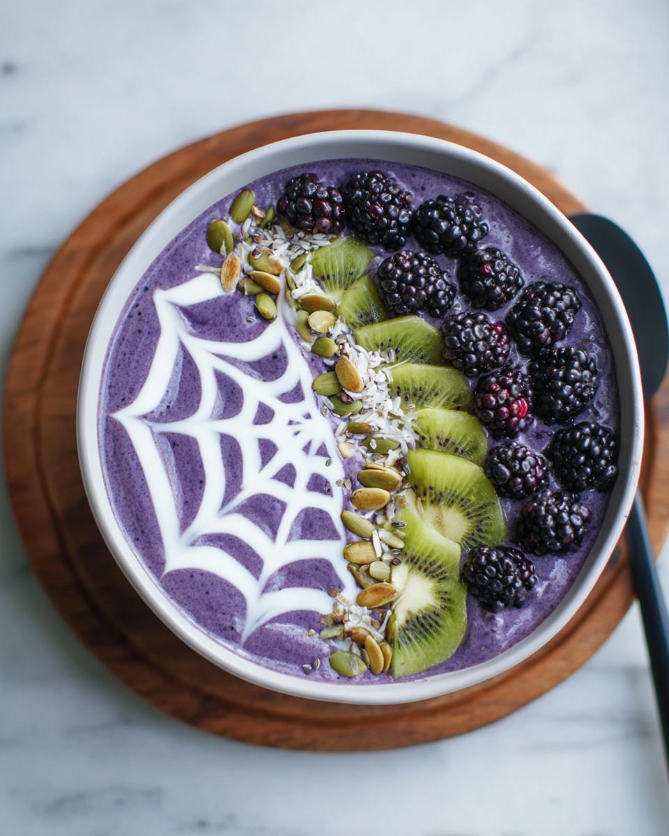 A vibrant purple Halloween smoothie bowl topped with blackberries, kiwi slices, seeds, and a white yogurt spiderweb.
