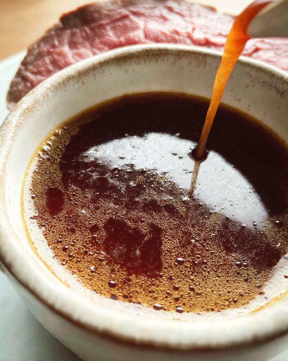 Close-up of rich, dark Homemade Au Jus without drippings being poured into a rustic bowl, with sliced roast beef visible in the background.