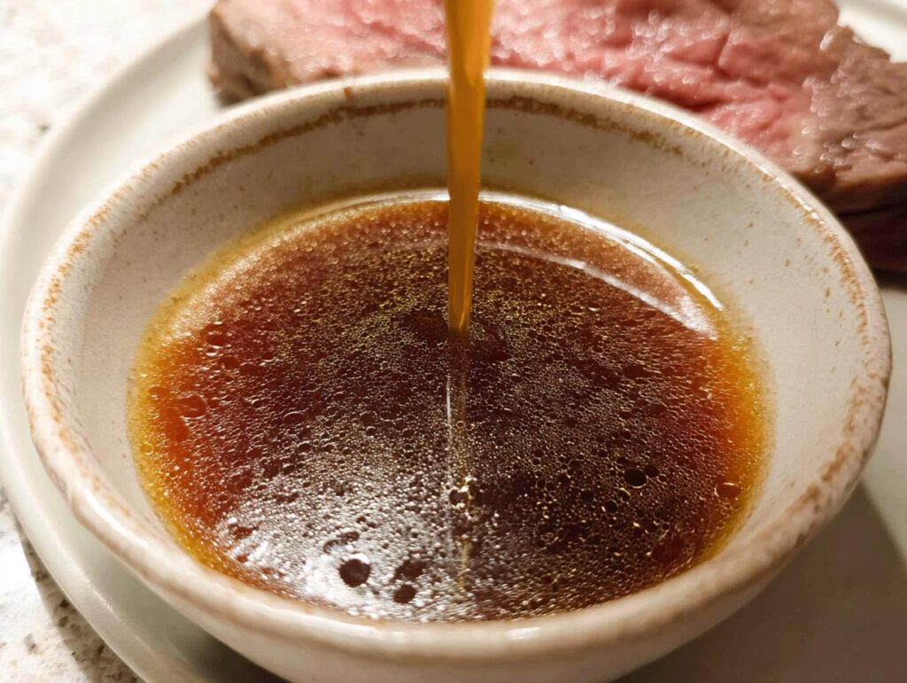 Close-up of rich, brown Homemade Au Jus without drippings being poured into a small ceramic bowl next to sliced roast beef.