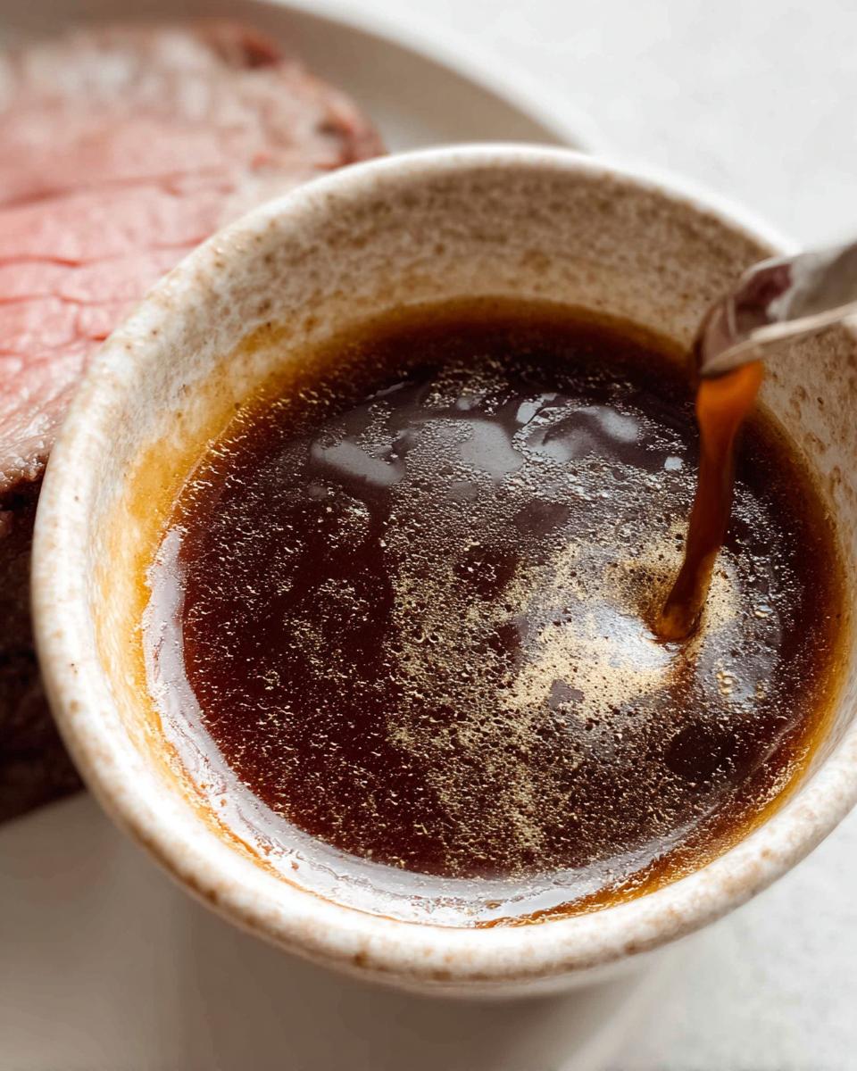 Close-up of rich brown Homemade Au Jus without drippings being poured into a small ceramic cup next to sliced roast beef.