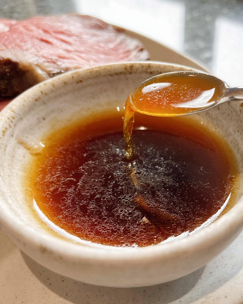 Close-up of a spoon dripping rich, brown Homemade Au Jus without drippings back into a small bowl, with roast beef in the background.