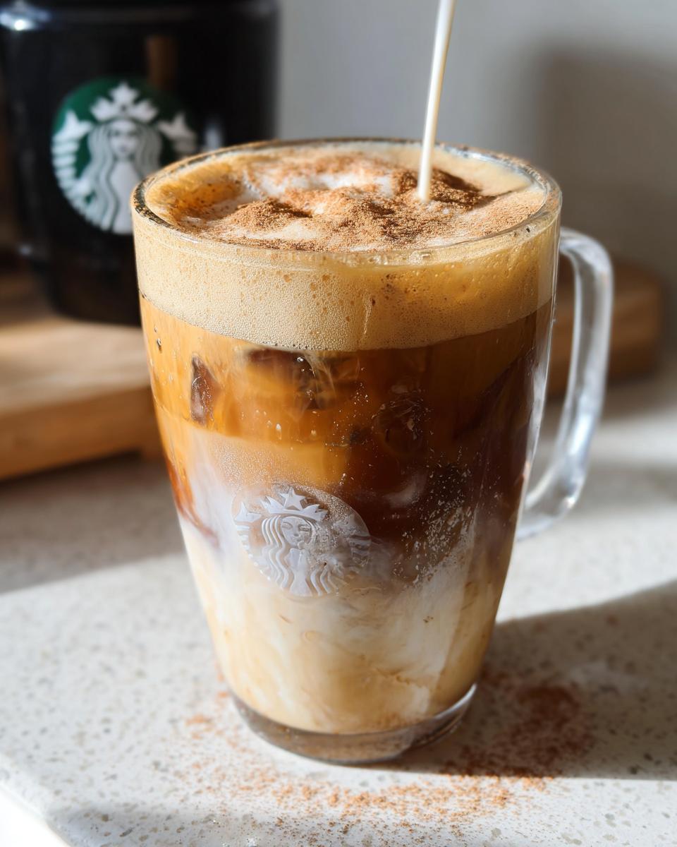 A glass mug of Iced Brown Sugar Oatmilk Shaken Espresso being finished with a stream of oat milk and cinnamon topping.