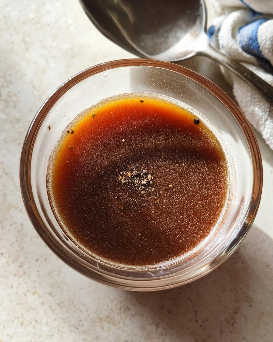Close-up of rich brown liquid au jus in a small glass bowl, topped with cracked pepper.