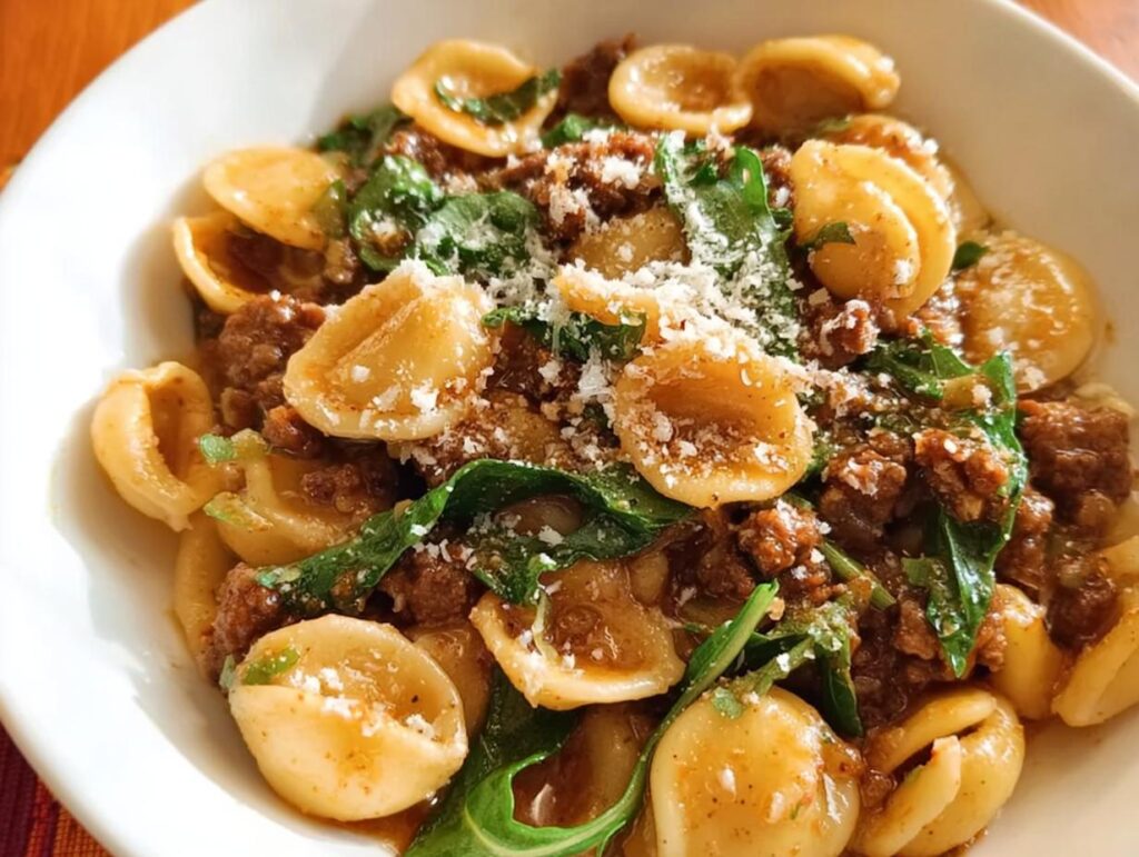 A close-up of a white bowl filled with orecchiette pasta, crumbled sausage, greens, and grated cheese. This is a dinner idea recipe everyone asks for.