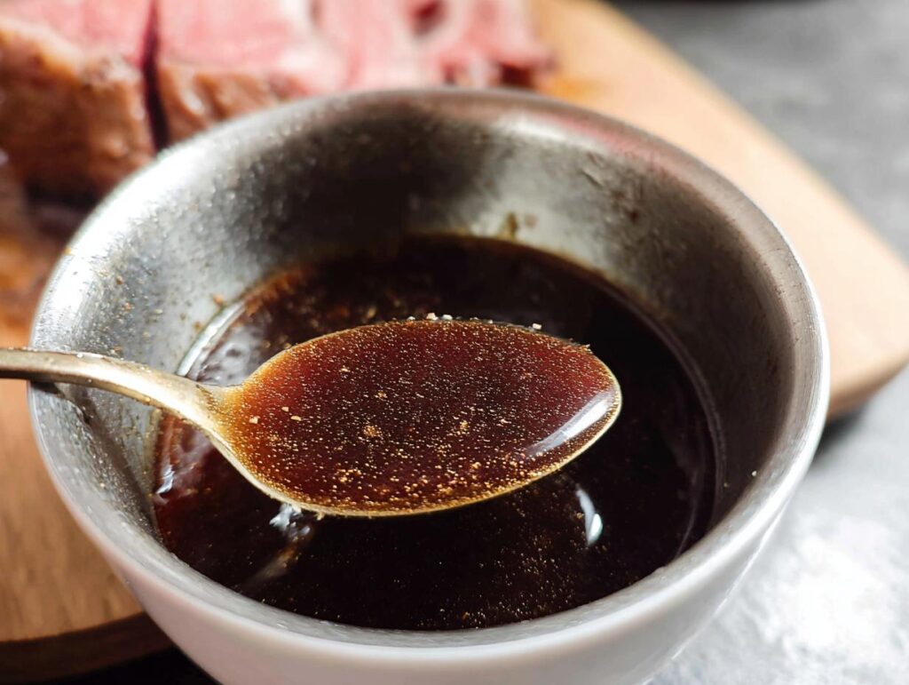 Close-up of a spoonful of rich, dark Prime Rib Au Jus Recipe sauce, with sliced roast beef visible in the background.
