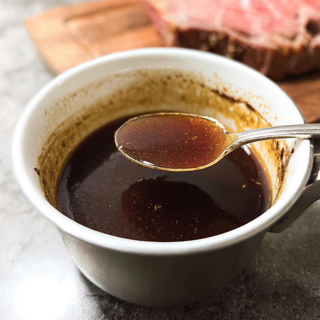 A spoonful of rich, dark brown au jus being lifted from a white mug, with prime rib visible in the background.