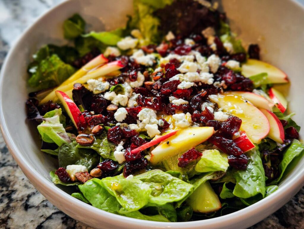 A vibrant Thanksgiving salad with mixed greens, apple slices, dried cranberries, blue cheese crumbles, and nuts.