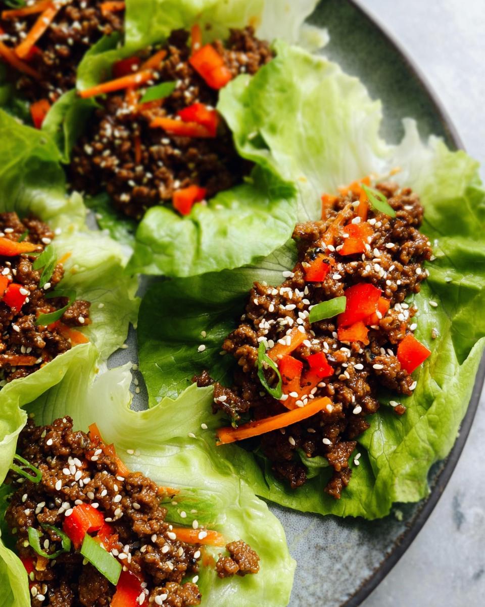 Close-up of three servings of Asian Ground Beef Lettuce Wraps filled with savory ground meat, topped with sesame seeds, red peppers, and scallions.