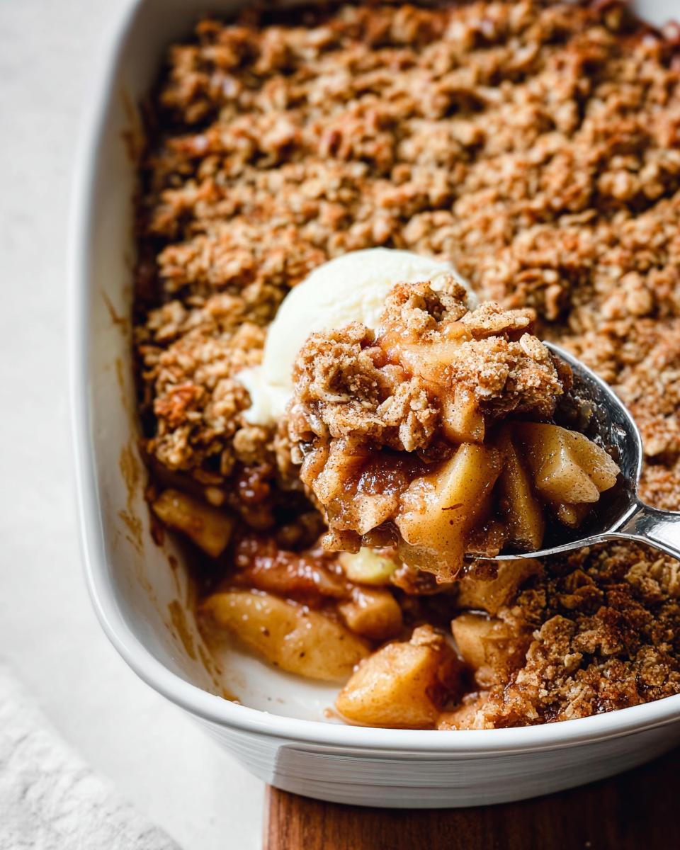 A spoonful of warm Classic Apple Crisp with Oat Crumble topped with melting vanilla ice cream being lifted from a white baking dish.