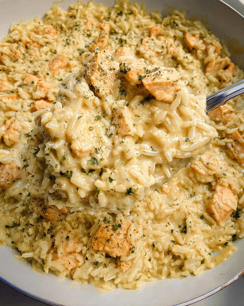 A spoonful of creamy chicken and rice skillet lifted from the pan, showing chunks of seasoned chicken.