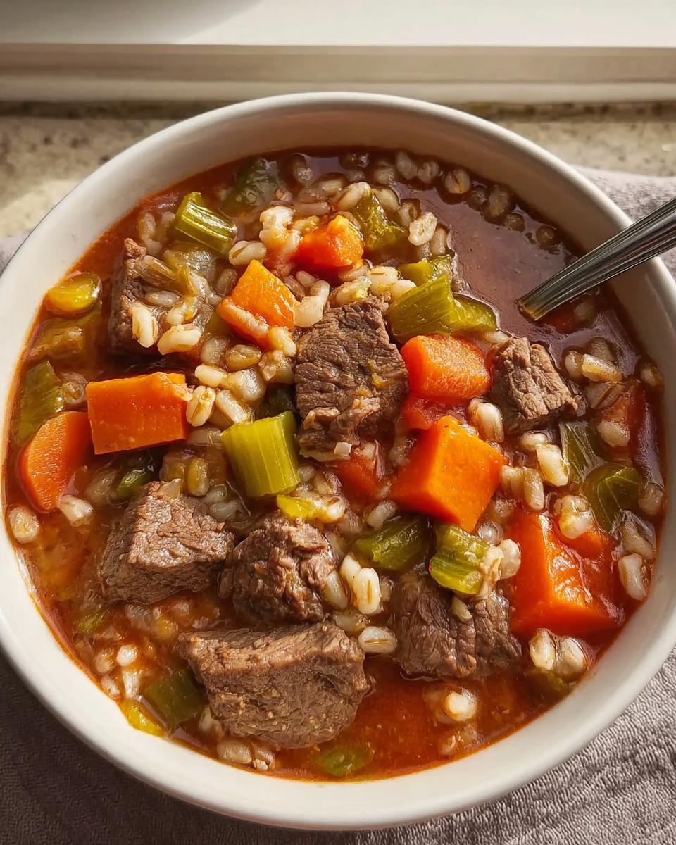 A close-up view of a bowl filled with Hearty Beef and Vegetable Barley Soup, showing chunks of beef, carrots, celery, and barley.
