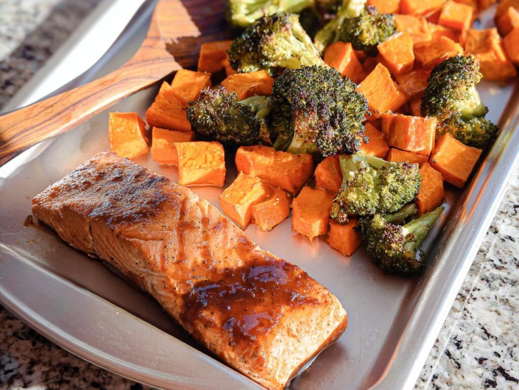 A cooked fillet of Maple Dijon Salmon Sheet Pan Dinner next to roasted broccoli and sweet potatoes on a baking sheet.