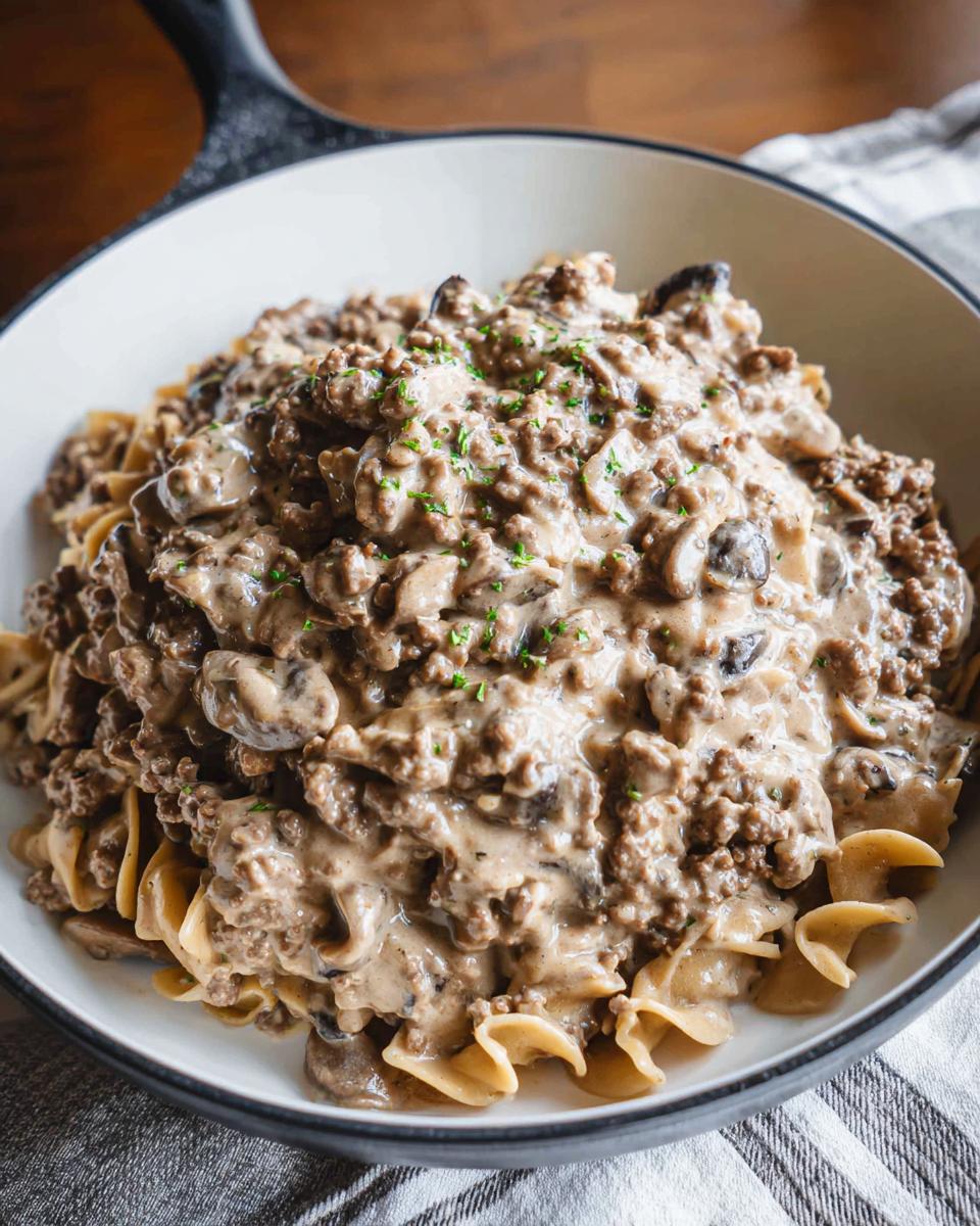 A close-up view of One Pot Ground Beef Stroganoff served over wide egg noodles in a white skillet, topped with parsley.