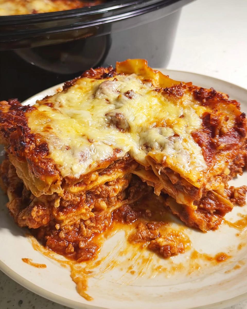 A generous slice of cheesy Slow Cooker Lasagna with Meat Sauce served on a plate, with the slow cooker visible in the background.