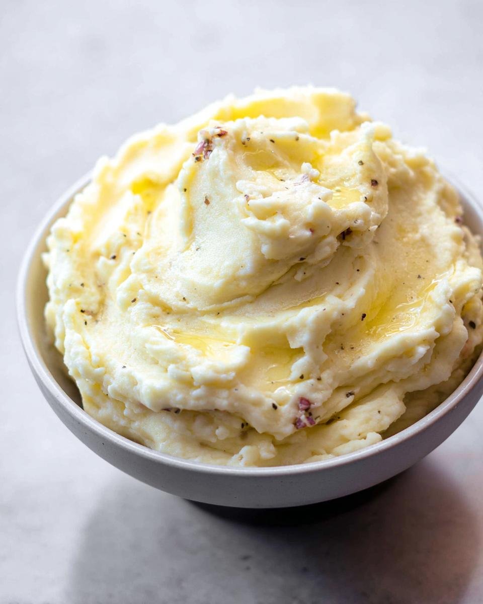 A close-up of a bowl filled with Ultra Creamy Garlic Mashed Potatoes, topped with melted butter and black pepper.