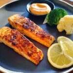 Two perfectly pan-seared salmon fillets served with steamed broccoli, cauliflower, a lemon slice, and dipping sauce.
