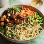 A vibrant bowl of 20-Ingredient Rice Bowl recipe featuring crispy tofu, broccoli, edamame, shredded carrots, and noodles, drizzled with sauce.
