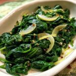 Close-up of a white dish filled with vibrant, wilted spinach, garnished with thinly sliced onions and red pepper flakes.