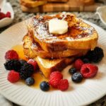 Stack of golden brown French toast topped with powdered sugar, butter, and syrup, garnished with fresh berries.