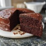 A slice of rich, moist chocolate cake with glossy chocolate frosting, part of a whole cake. A perfect example of 7-Ingredient Cake Ideas.