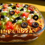 A vibrant 7 layer taco dip, perfect for Taco Tuesday recipes, topped with shredded cheese, olives, tomatoes, lettuce, and onions.