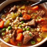 A close-up of a bowl of hearty beef barley soup, featuring tender beef chunks, barley, carrots, peas, and celery in a rich broth.