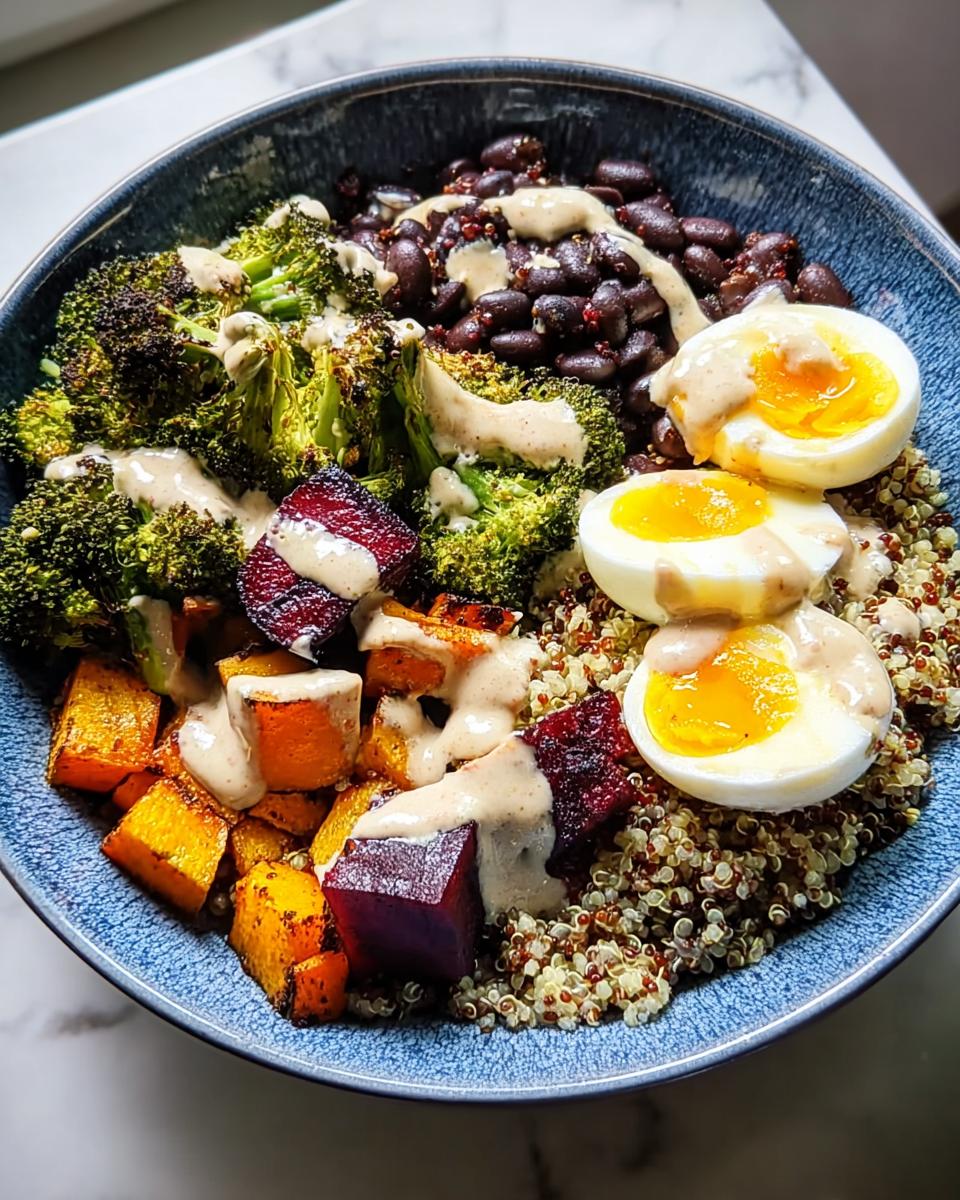 A vibrant breakfast bowl featuring quinoa, black beans, roasted broccoli, butternut squash, beets, and halved hard-boiled eggs, drizzled with a creamy sauce.