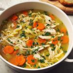 A close-up of a steaming bowl of homemade chicken noodle soup, featuring shredded chicken, egg noodles, carrots, and celery, garnished with parsley.