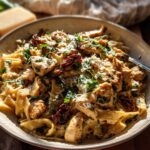A close-up of a pasta bowl filled with fettuccine, chicken pieces, sun-dried tomatoes, and fresh herbs, topped with grated cheese.