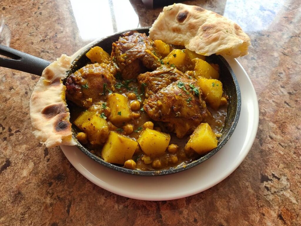 A close-up of a cast-iron skillet filled with a rich chicken thigh recipe, featuring tender chicken pieces, golden potatoes, and chickpeas in a savory sauce, garnished with parsley and served with flatbread.