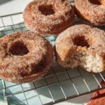 Close-up of homemade cinnamon sugar donuts on a cooling rack, one with a bite taken out, showcasing a fluffy interior.