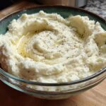 A close-up of creamy mashed potatoes in a bowl, topped with melted butter and herbs.