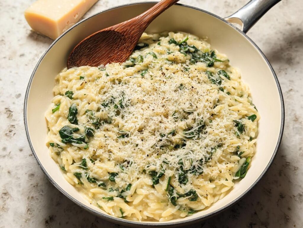 Close-up of creamy spinach orzo in a pan, topped with grated parmesan cheese and black pepper.