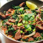 A close-up of a bowl filled with an easy beef and spinach stir-fry, garnished with lime and scallions.