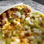 A close-up of a breakfast burrito filled with scrambled eggs, sausage, and onions, topped with diced celery and spices.