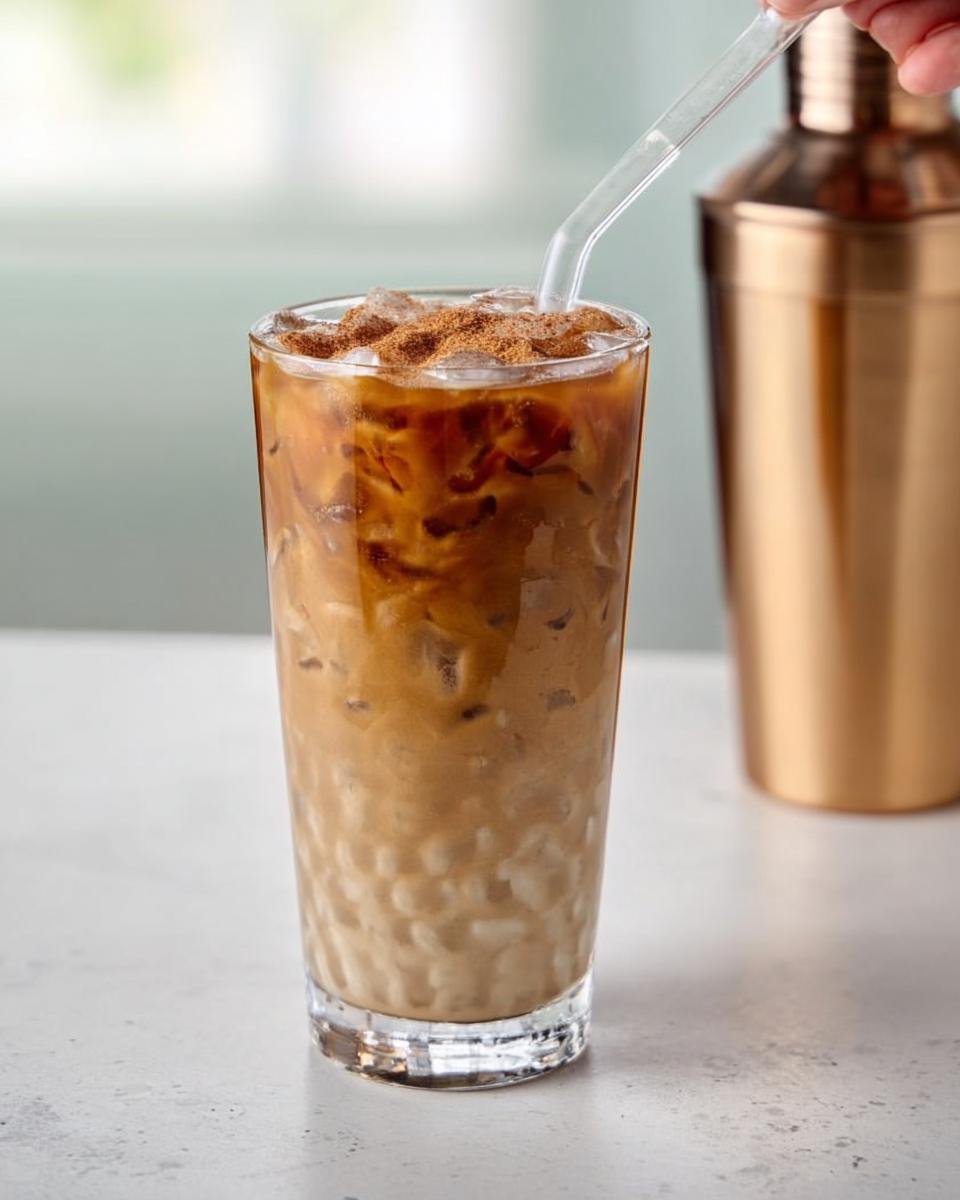Close-up of a tall glass filled with iced coffee, milk, and cinnamon, a straw inserted. Part of a gold shaker in the background.