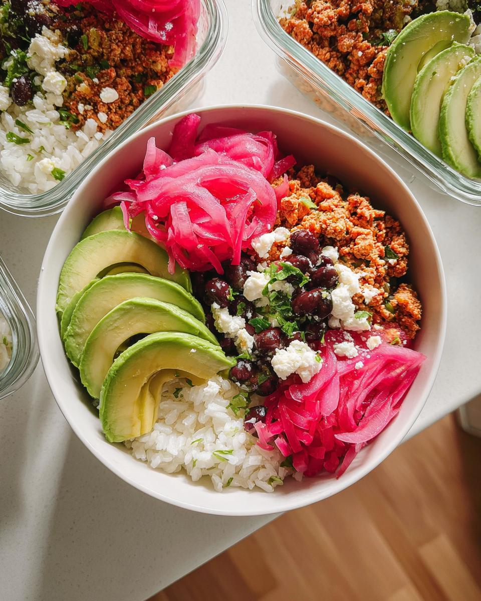 A colorful and vibrant bowl of rice, seasoned ground meat, black beans, feta cheese, pickled red onions, and sliced avocado for meal prep.