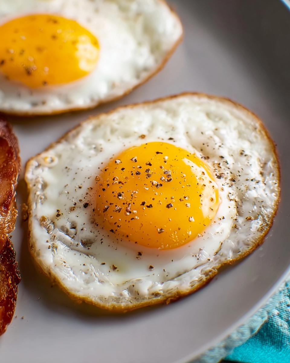 Close-up of two perfectly fried eggs with runny yolks, seasoned with black pepper, a favorite egg recipe.