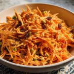 A close-up of a bowl filled with shredded carrot salad, mixed with raisins and green peas, perfect for veggie sides recipes.
