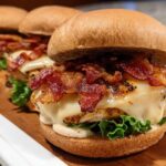 Three restaurant-style chicken bacon burgers with melted cheese and lettuce, served on a wooden board.