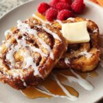 Two pieces of cinnamon roll French toast drizzled with icing and syrup, topped with butter and raspberries. Restaurant-Style Breakfast Ideas Recipes at Home.